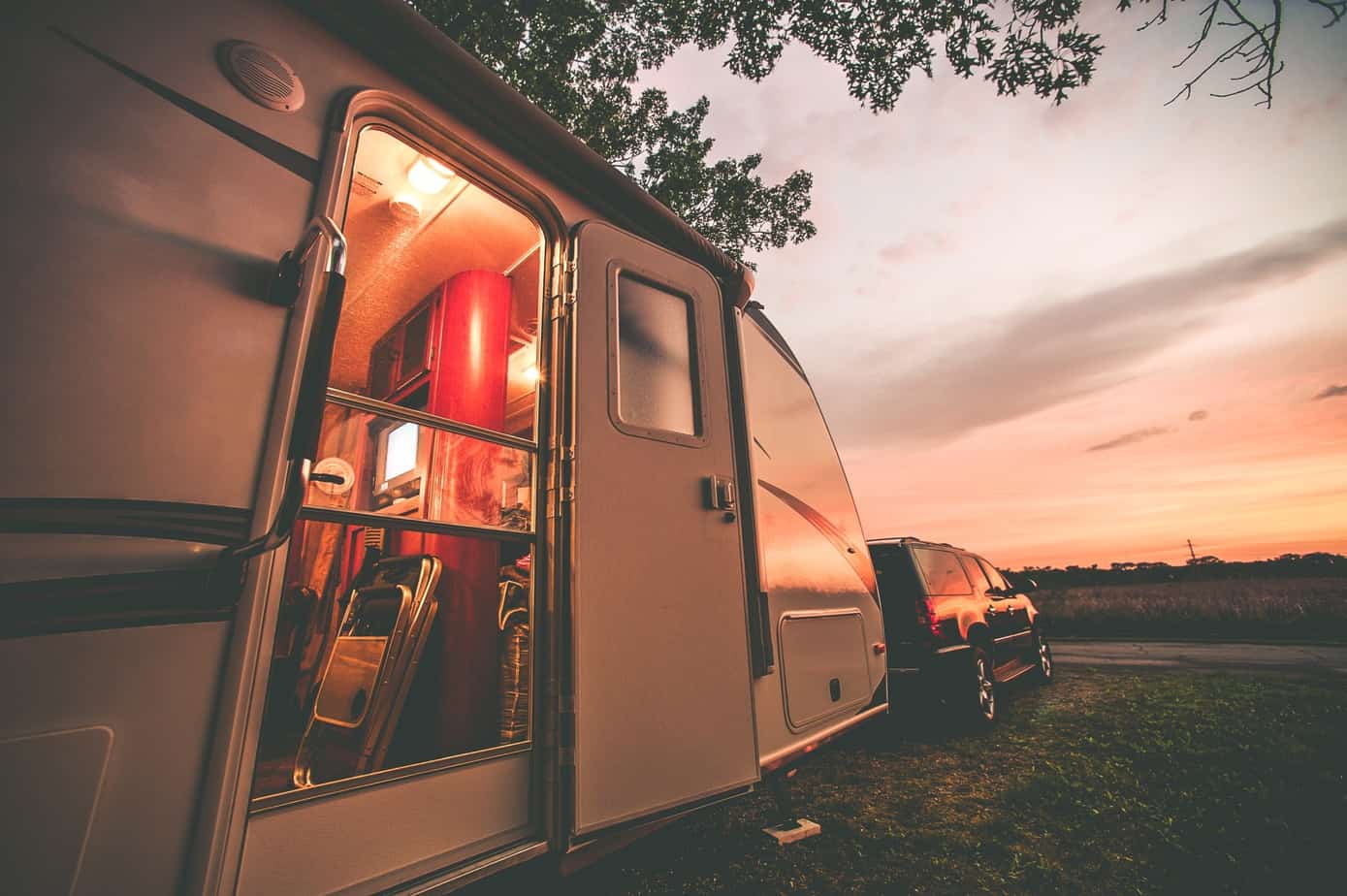 An RV is attached to a black car with its door open as the sky turns purple at night.