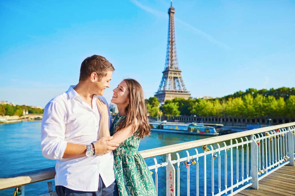 Young couple in love stands on a bridge by the river with the Eiffel Tower behind them.