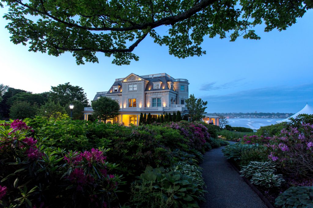 A big white estate at night near the water. Green gardens are in the front.