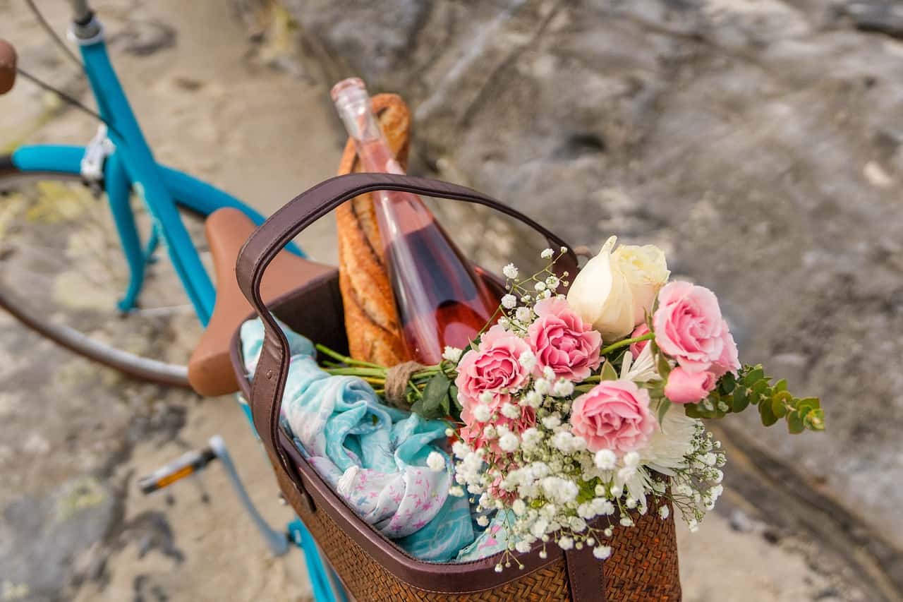 Picnic basket on the back of a bike is filled with bread, wine, and flowers.