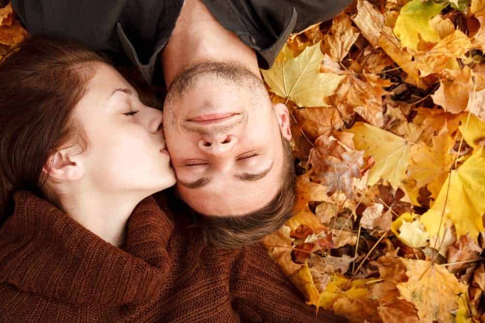 A couple is laying on fall leaves as the girl kisses the boy's cheek.