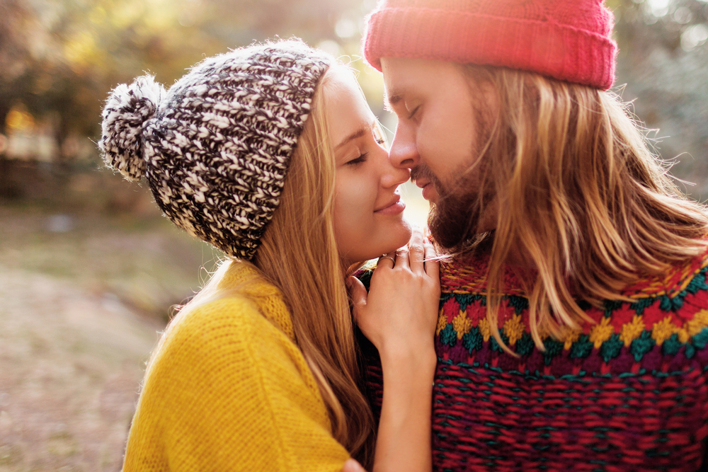 A couple in brightly colored sweaters and winter beanies rub noses outdoors in the late sun.