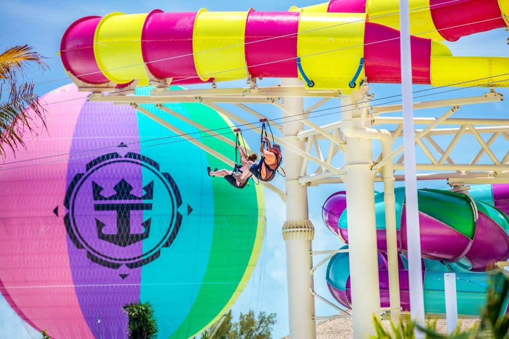 Two riders zip lining in front of a colorful slide and hot air balloon. 