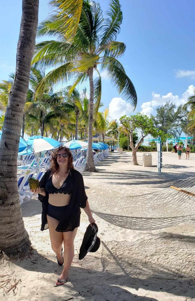 A woman stands on the beach smiling surrounded by palm trees and an empty hammock.