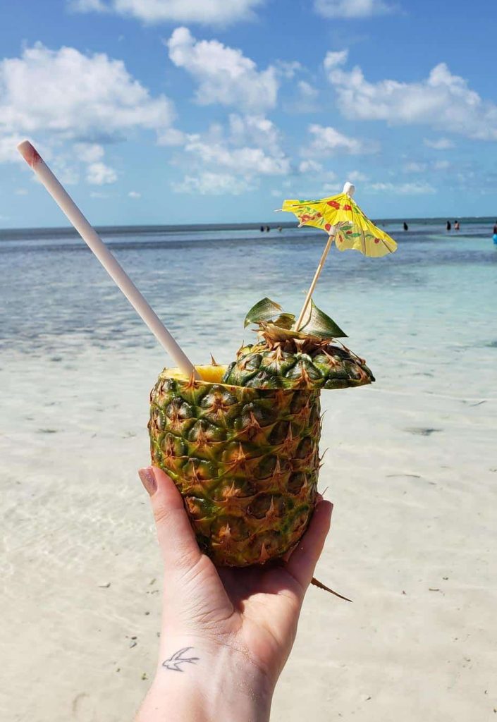 A person holds a pineapple filled with a drink with the ocean behind.