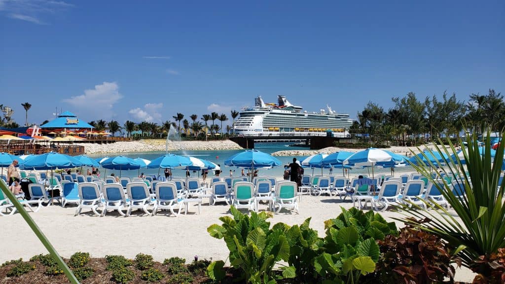 People relax in blue chairs by the ocean in white sand. A cruise ship is seen in the distance.