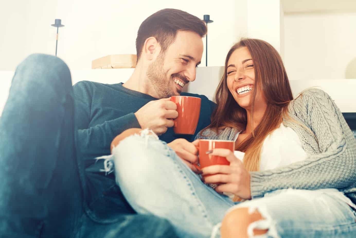 A couple holds red mugs and laughs together on a couch.