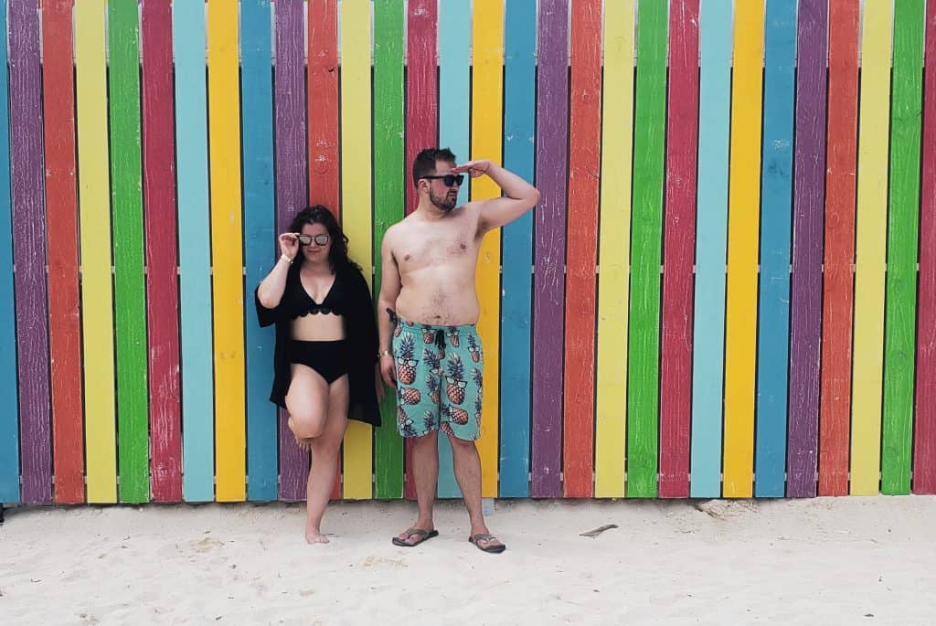 A couple in swimsuits smiles in front of a rainbow fence.