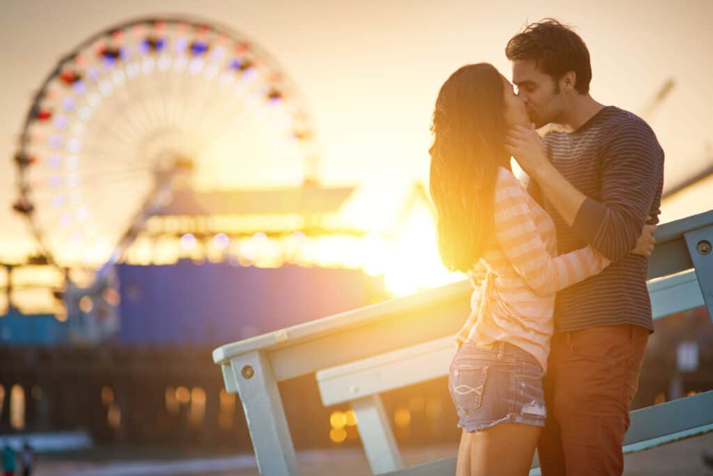A couple kisses outdoors with a Ferris wheel in the distance.