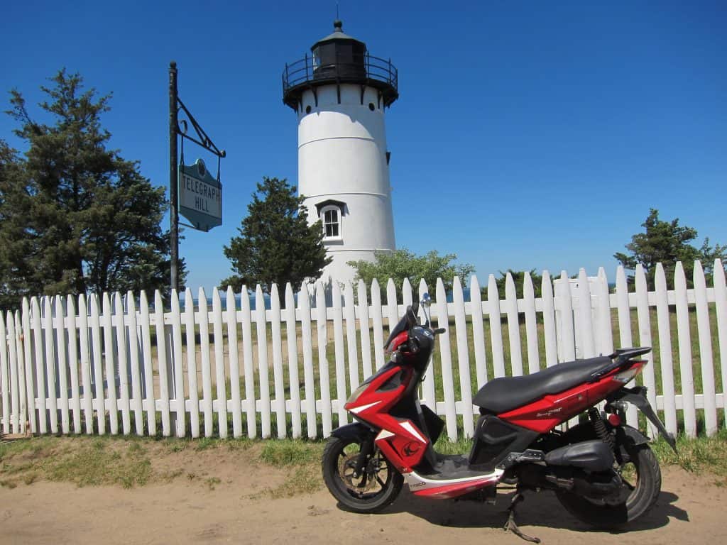 Red moped is parked in front of a white fence and lighthouse.