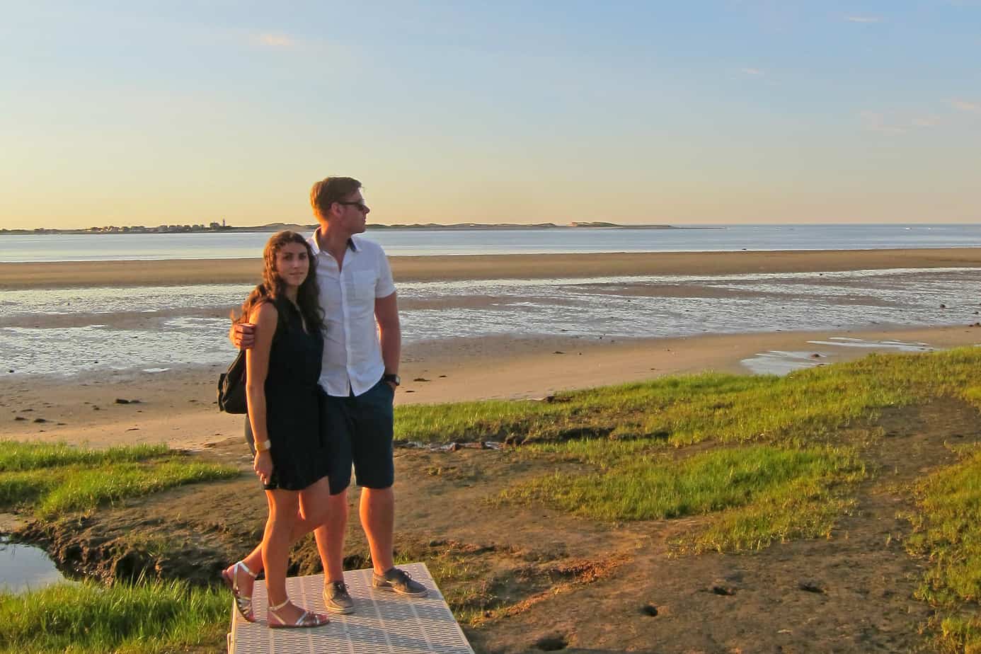 A couple stands smiling on the beach with water behind.