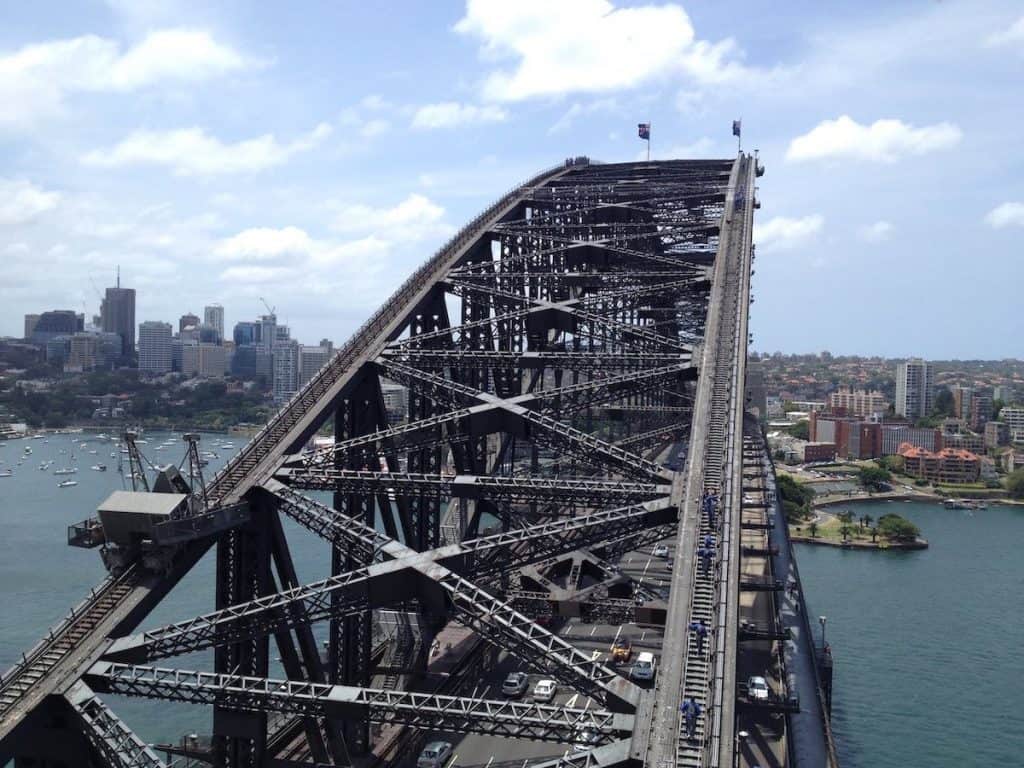 People walking up and over a bridge with cars driving on it. A city skyline is in the ditsance.