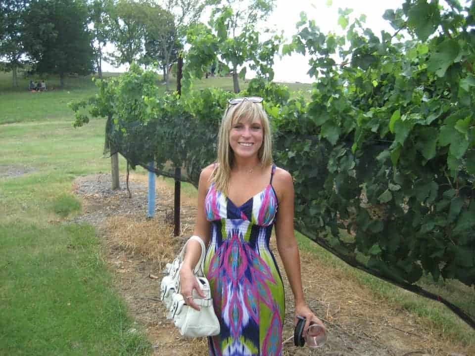 Woman in a pink/green/blue dress holding a glass of wine in a vineyard.