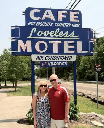 A couple smiles in front of a blue sign that says Cafe Hot Biscuits Country Ham Loveless Motel Air Conditioned.
