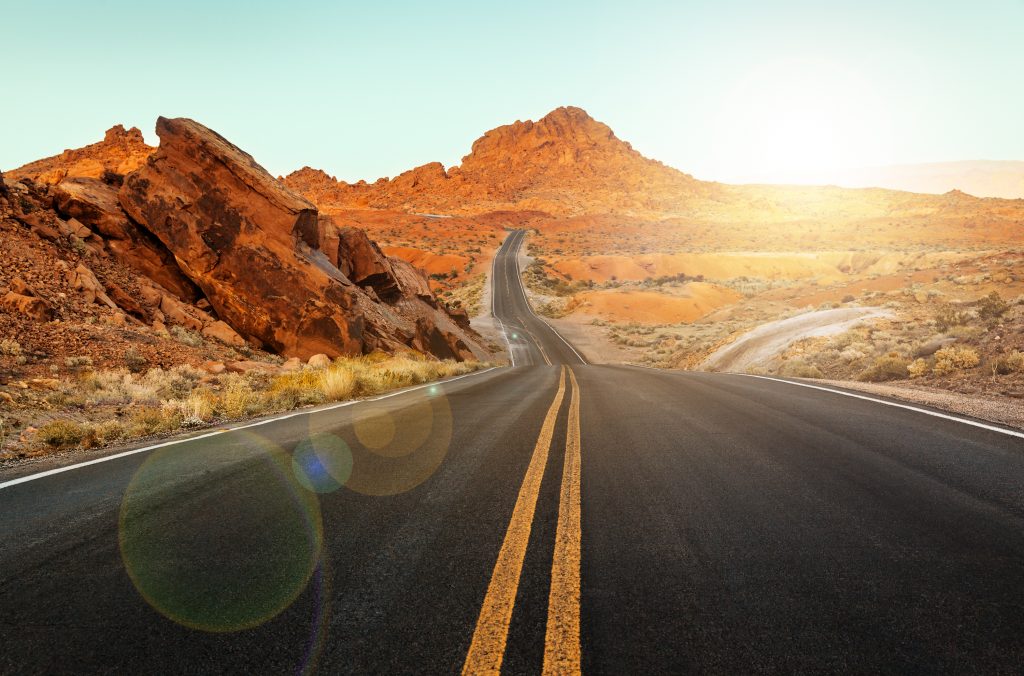 A wide open road surrounded by red desert formations as the sun glows.