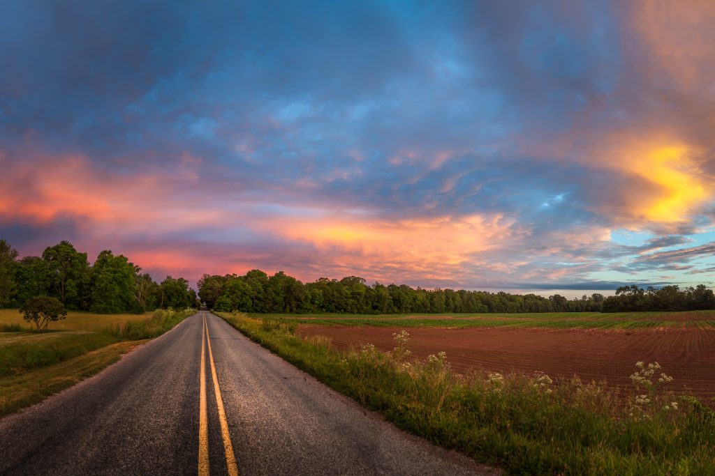 An empty country road is lined by trees under a purple sky.