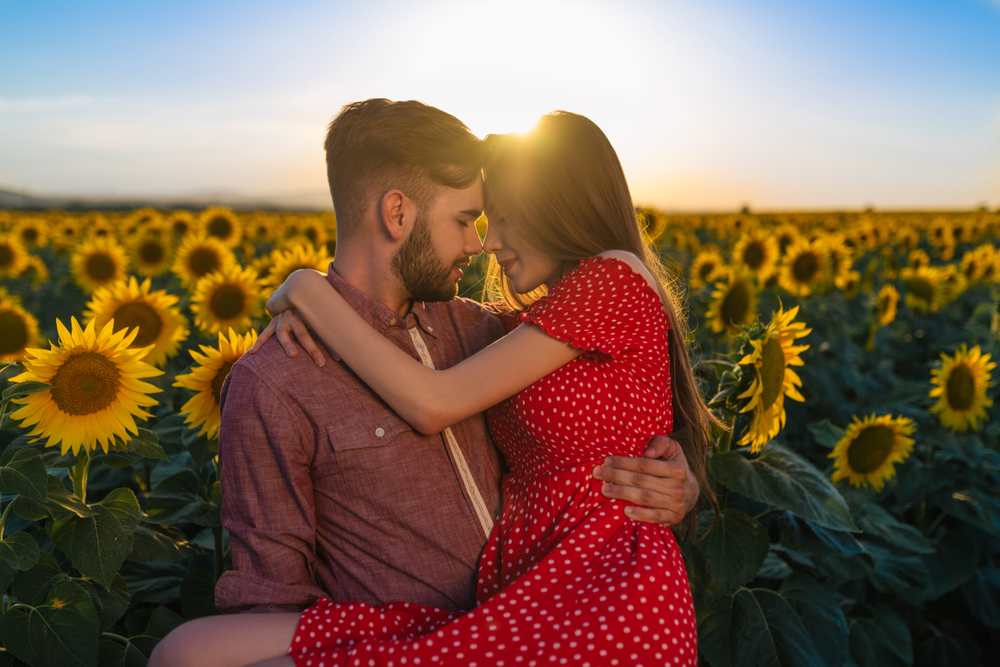 A couple embraces in front of a sunflower field. The man holds the woman in his arms and she wears a red dress.