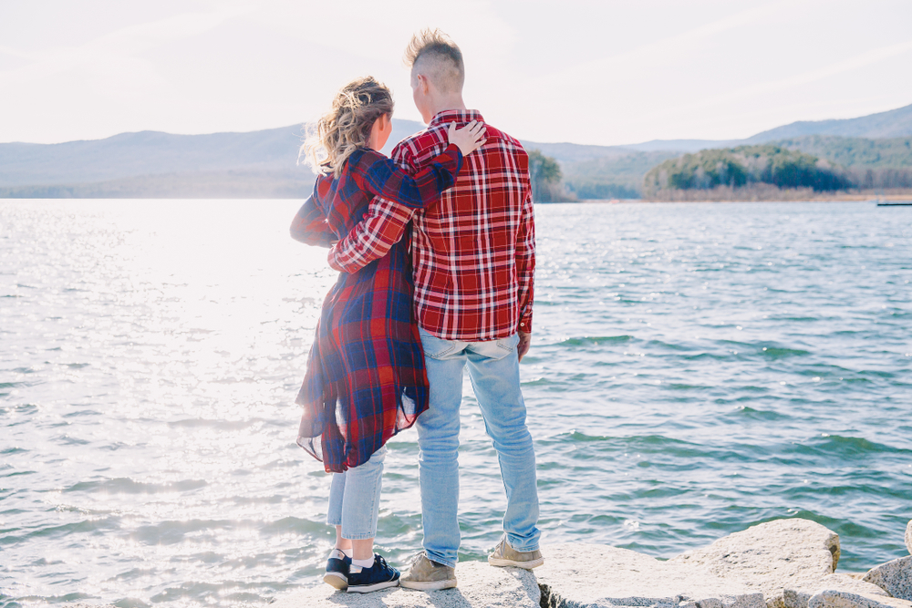 A couple stands in front of a lake with their backs to their camera. They have their arms on each other\'s shoulders. They both wear red and blue plaid shirts.