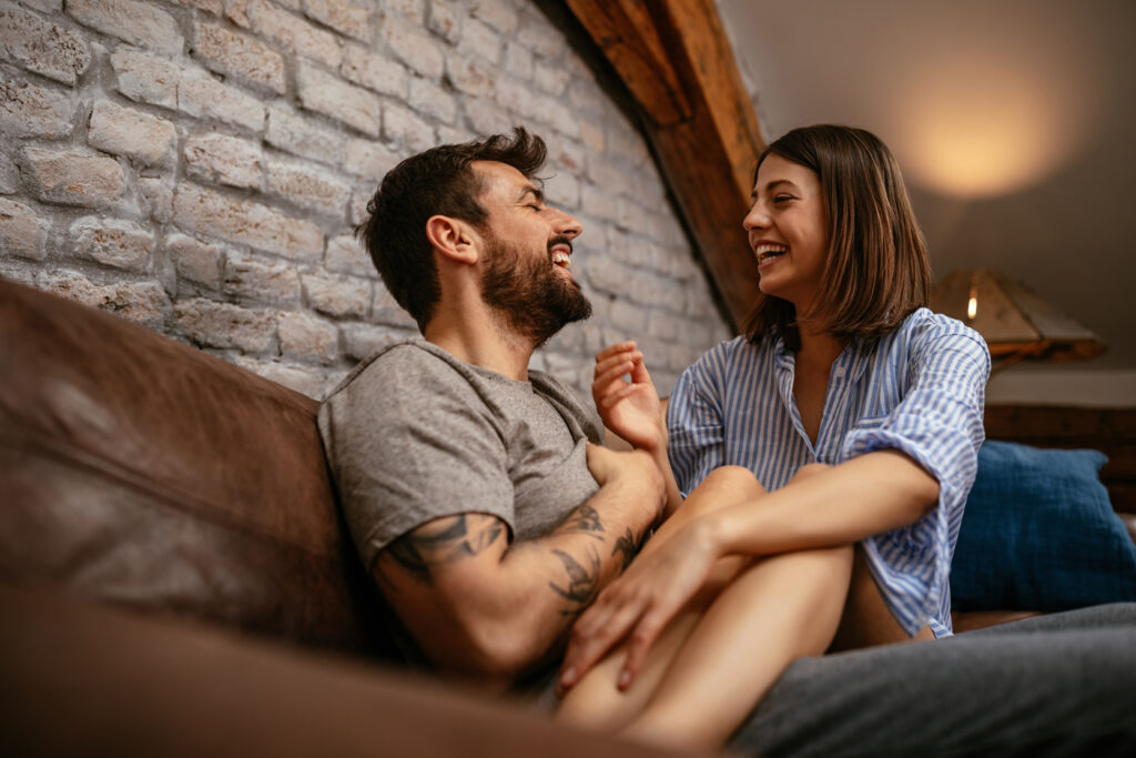 A couple laughs with one another while on a brown leather couch.