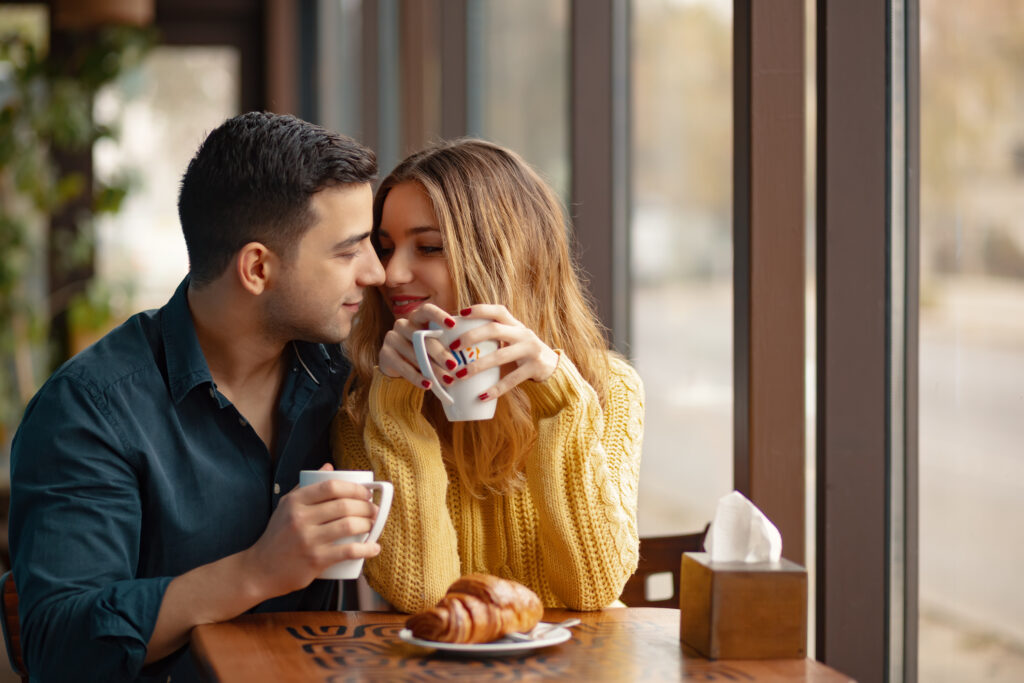 Young couple sitting at a table on a date in a coffee shop while drinking coffee. A croissant is on a plate in front of them.