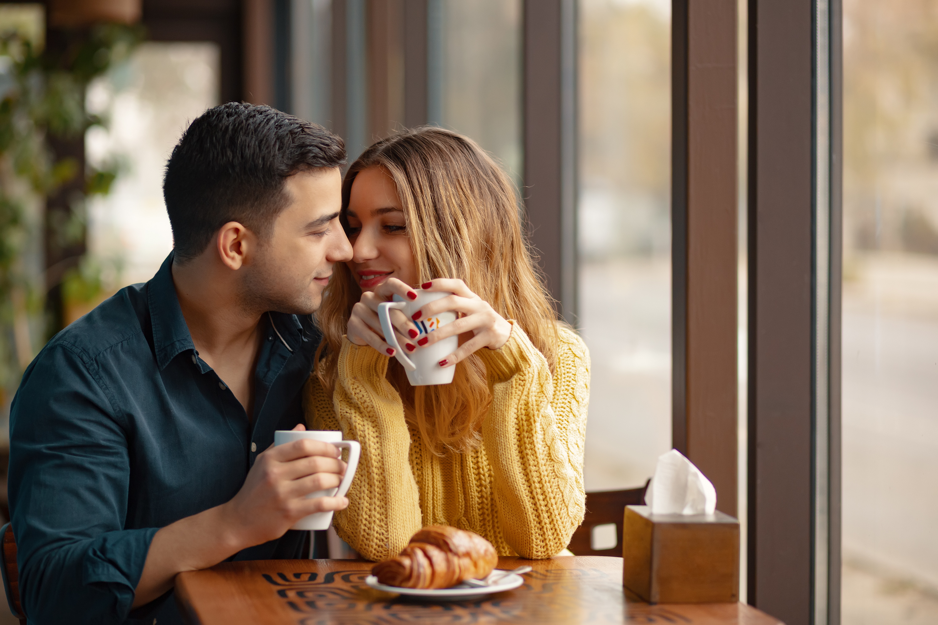 A young couple sitting at a table in a coffee shop while drinking coffee, with a croissant on a table in front of them.