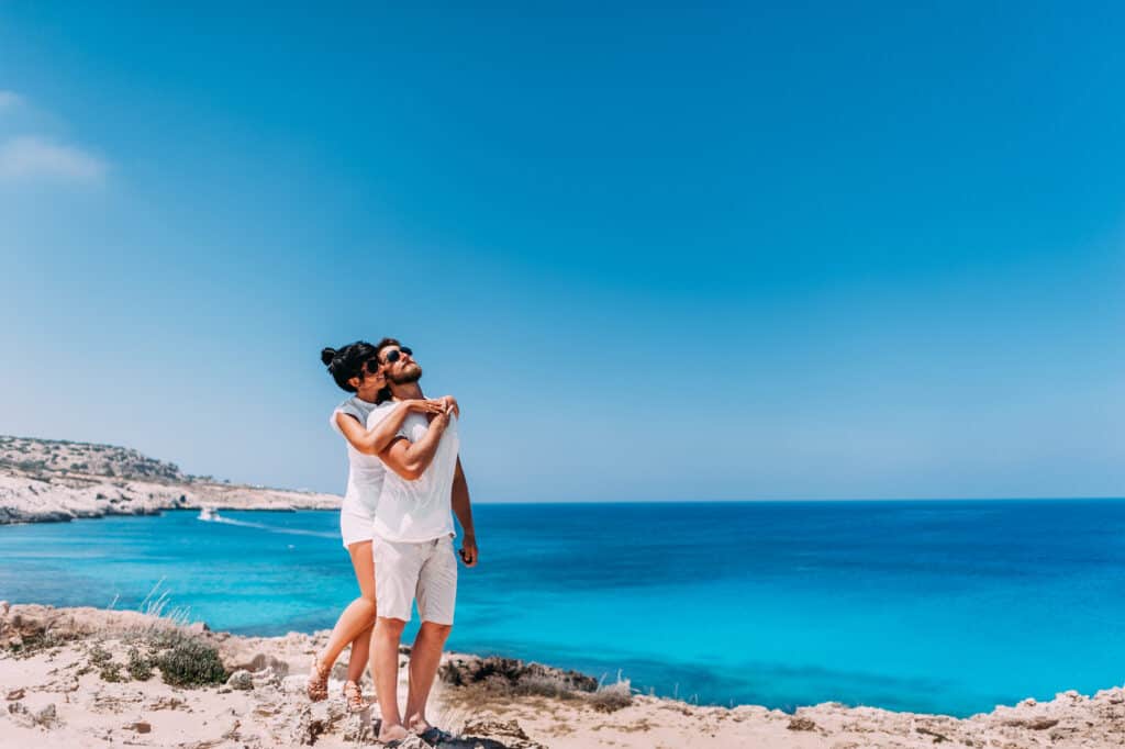 A happy couple by the sea hugs one another under a blue sky.