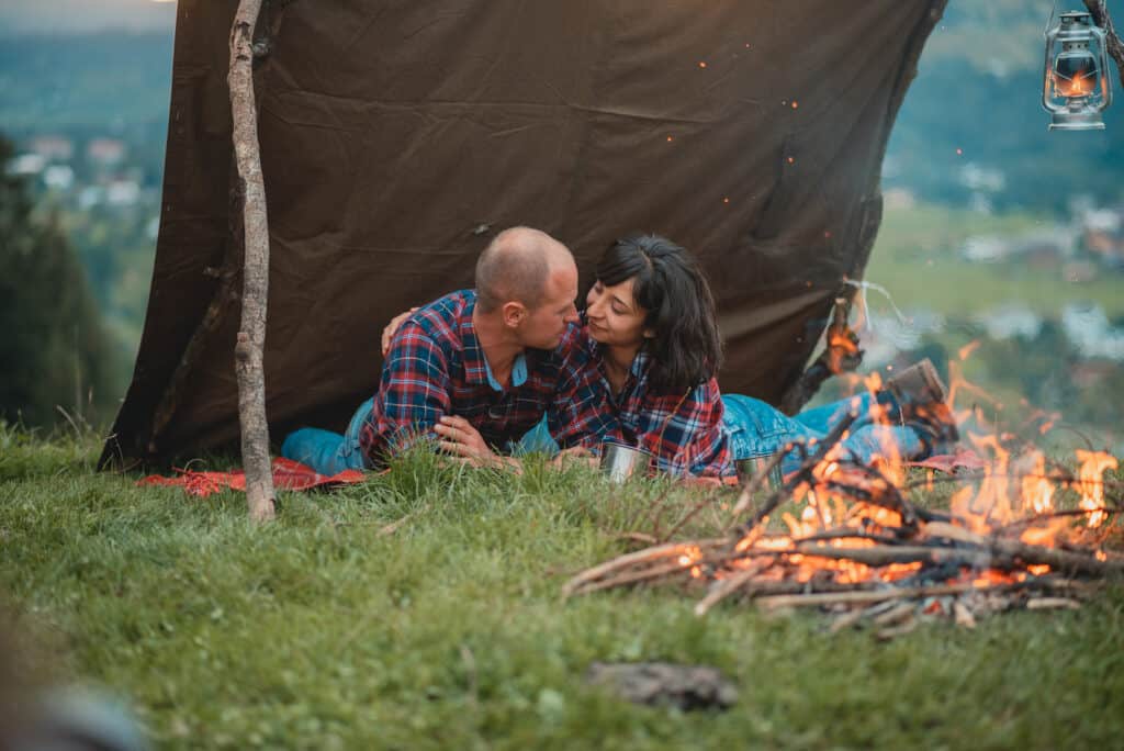 A couple lays outdoors in front of a fire under a brown tarp.
