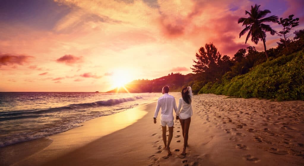 A couple holds hands while walking along the beach at sunset.