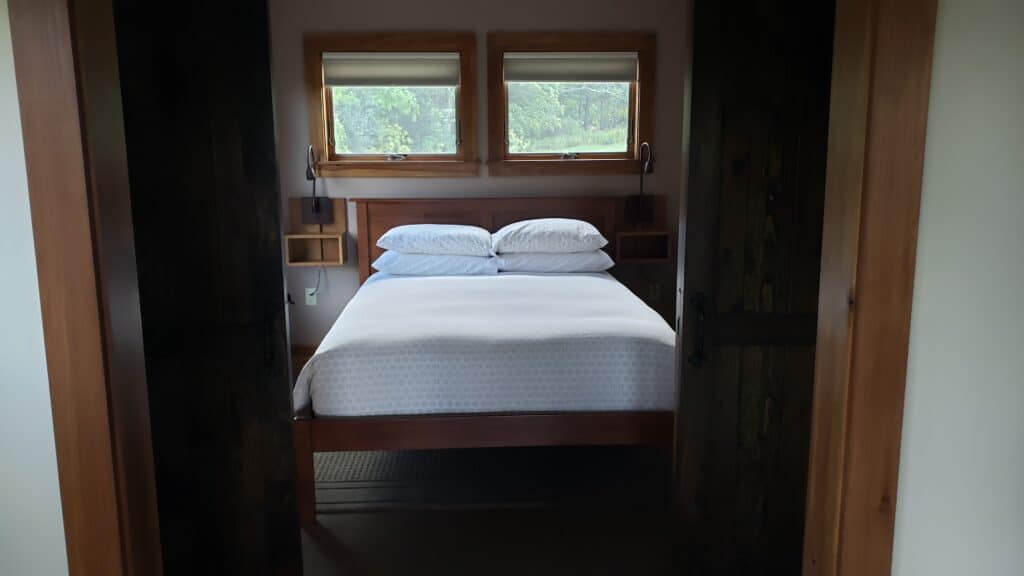 View through a doorway of a perfectly-made bed with white bedding. Two windows are side by side above the bed frame.