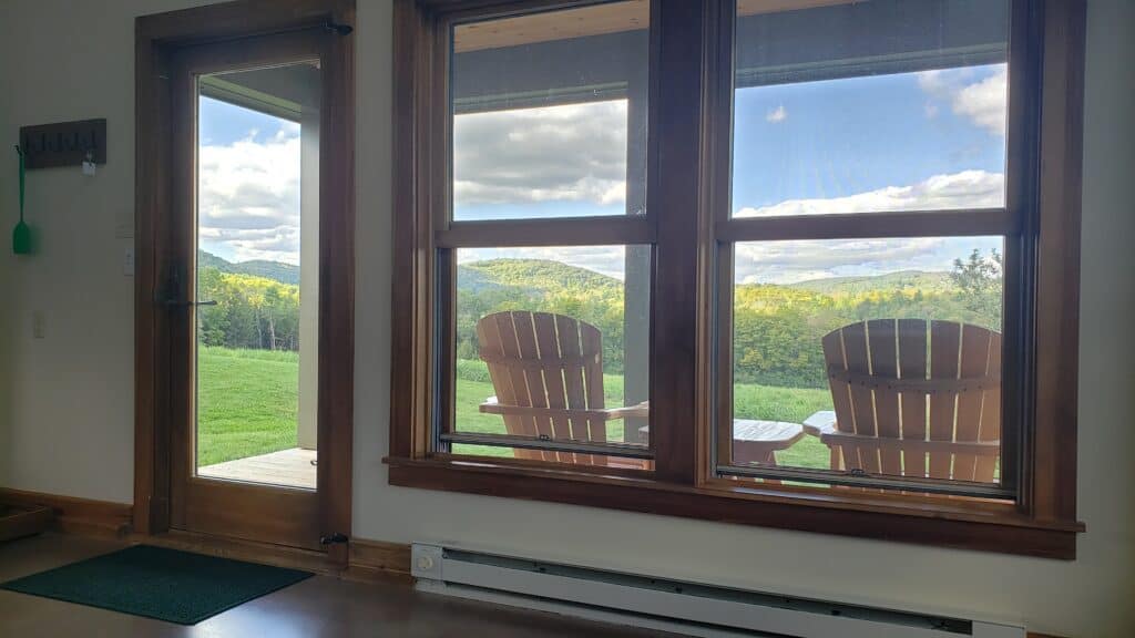 View through double windows of two chairs sitting on a porch. Rolling mountain landscape can be seen in the distance under a blue cloudy sky.