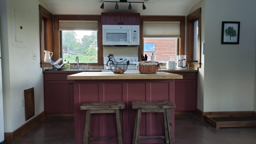 Kitchen with maroon cabinetry. Two baskets sit on the kitchen island, and there\'s a view of greenery out the two windows.