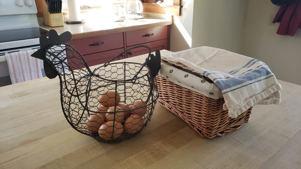 A wicker basket with a cloth on top sits next to a wire hen-shaped basket filled with eggs on a kitchen counter.