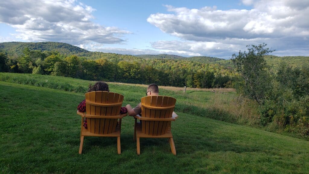 Man and woman sitting outdoors looking at the mountains in the distance under a blue cloudy sky. They are sitting in yellow outdoorsy chairs.