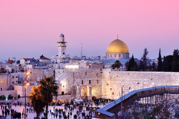 People walk around tall brick walls next to a church under a pink sky.