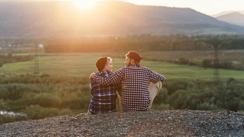 A young couple sits at the top of a mountain and looks at each other at sunset.