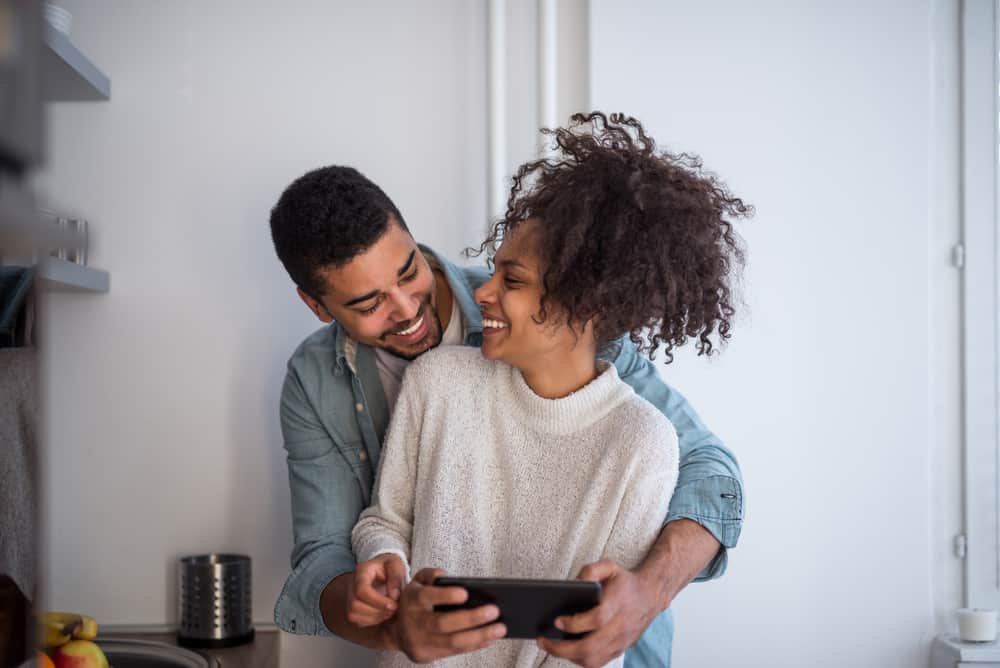 A couple laughs with their arms around each other while seeing a video on a phone.