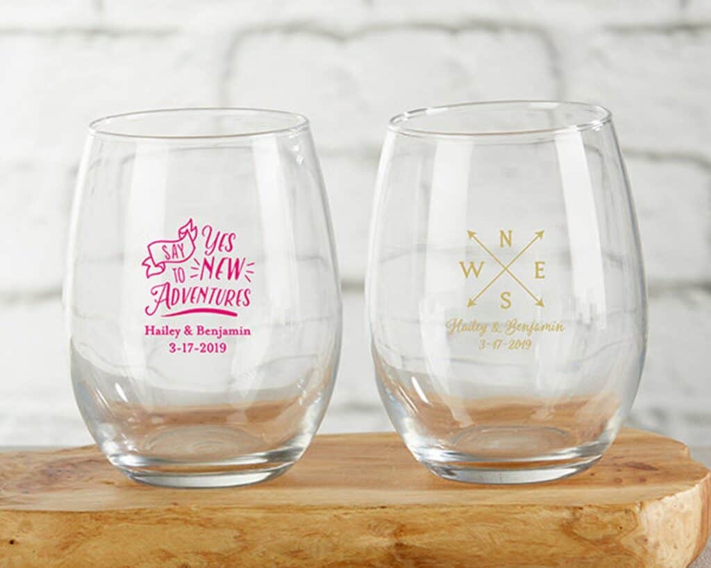 Two wine tumblers are empty. A pink one says Say yes to new adventures with the couples name and wedding date underneath. The other has grey writing with a compass.