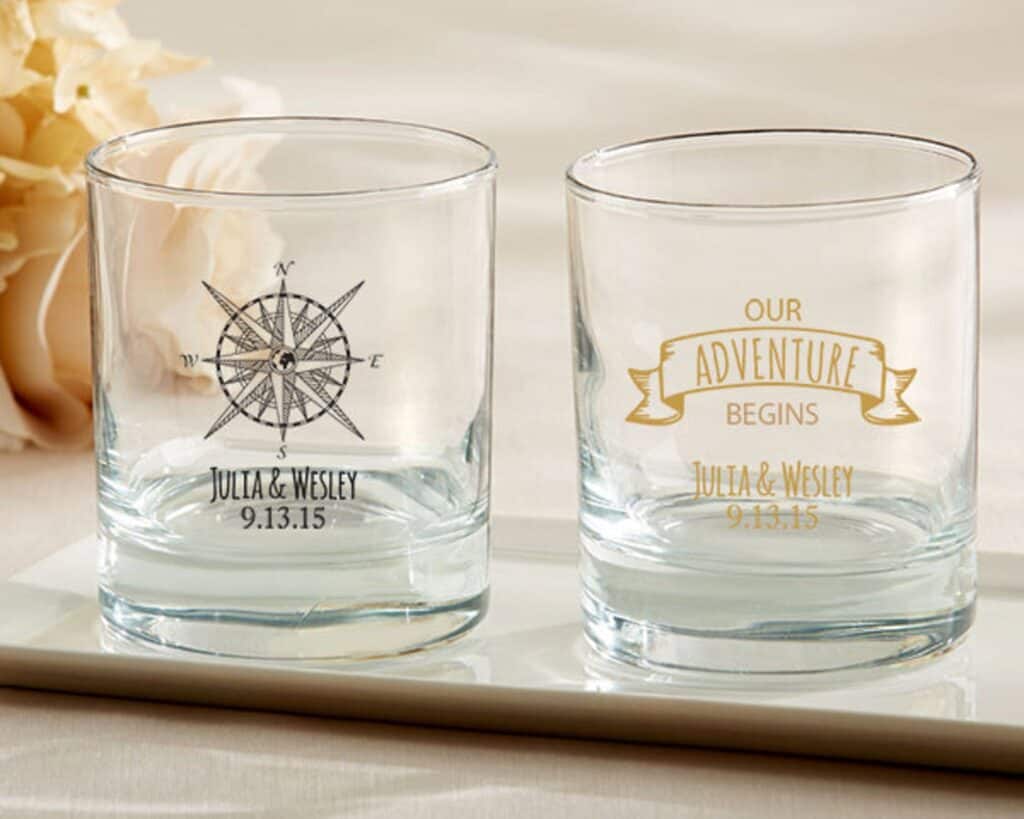 Two whiskey glasses are empty. One has a compass with a couple\'s name and wedding date. The other says Our Adventure Begins.