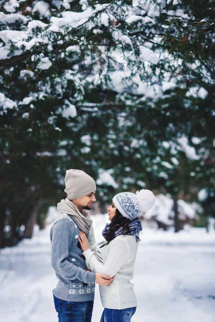 A couple laughs and smiles at one another surrounded by a snowy forest.