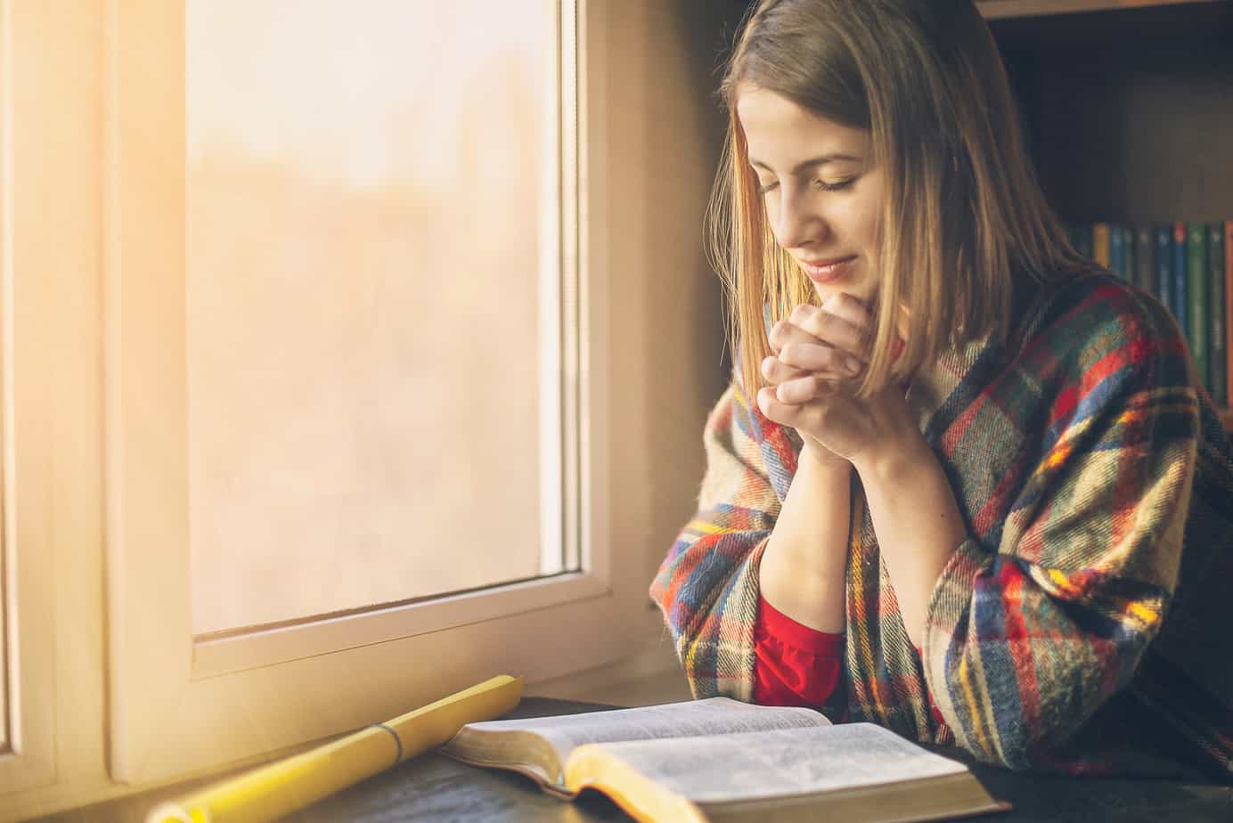 A woman has her hands grasped while reading a bible near the window.