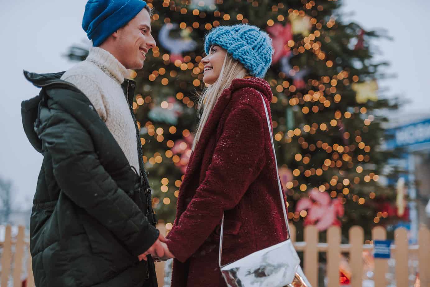 A couple holds hands while smiling at each other, with blurred our Christmas trees behind them.