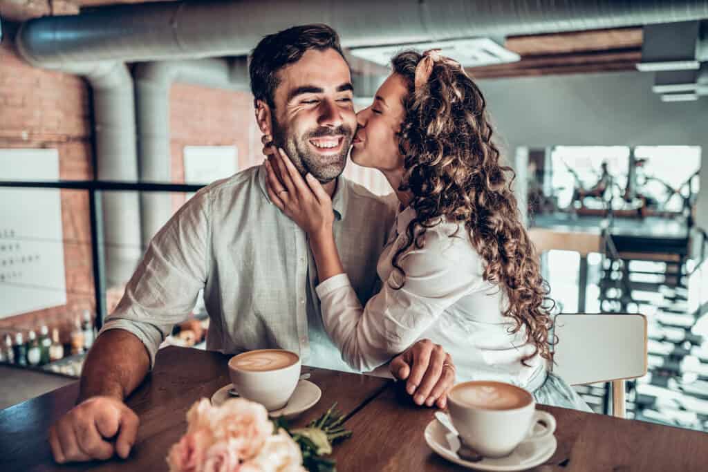 A woman kisses a man\'s cheek in front of coffee cups.