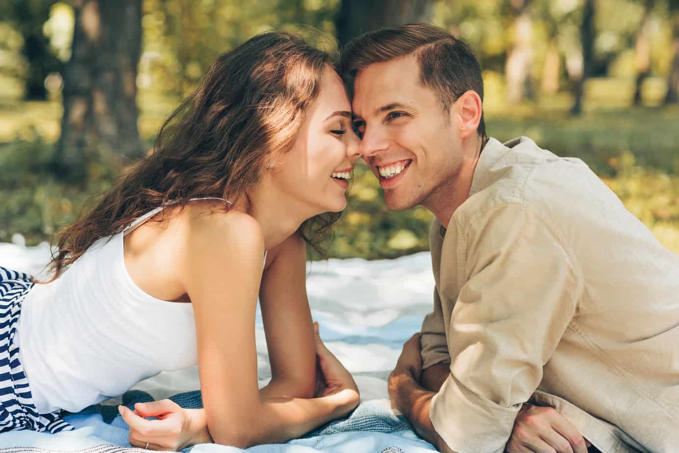 A couple sits nose to nose outdoors on a picnic blanket.