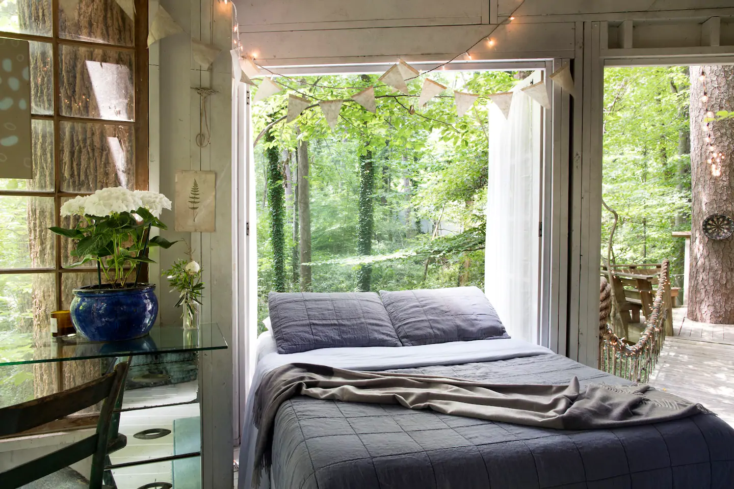 A large grey bed in a bedroom with a view of greenery right out the window.
