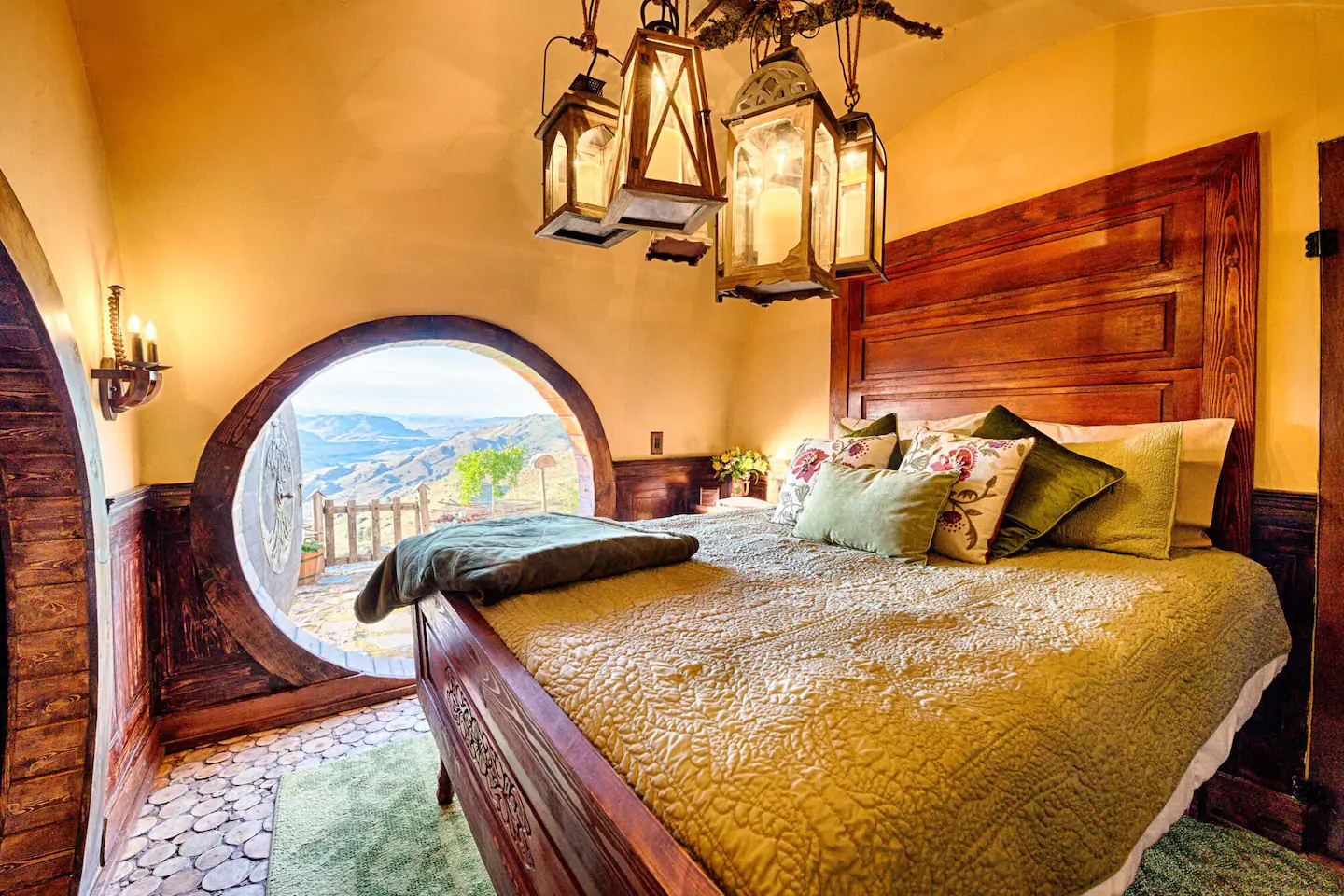 A bed with yellow bedding is made with green throw pillows. A circle door leads outside.