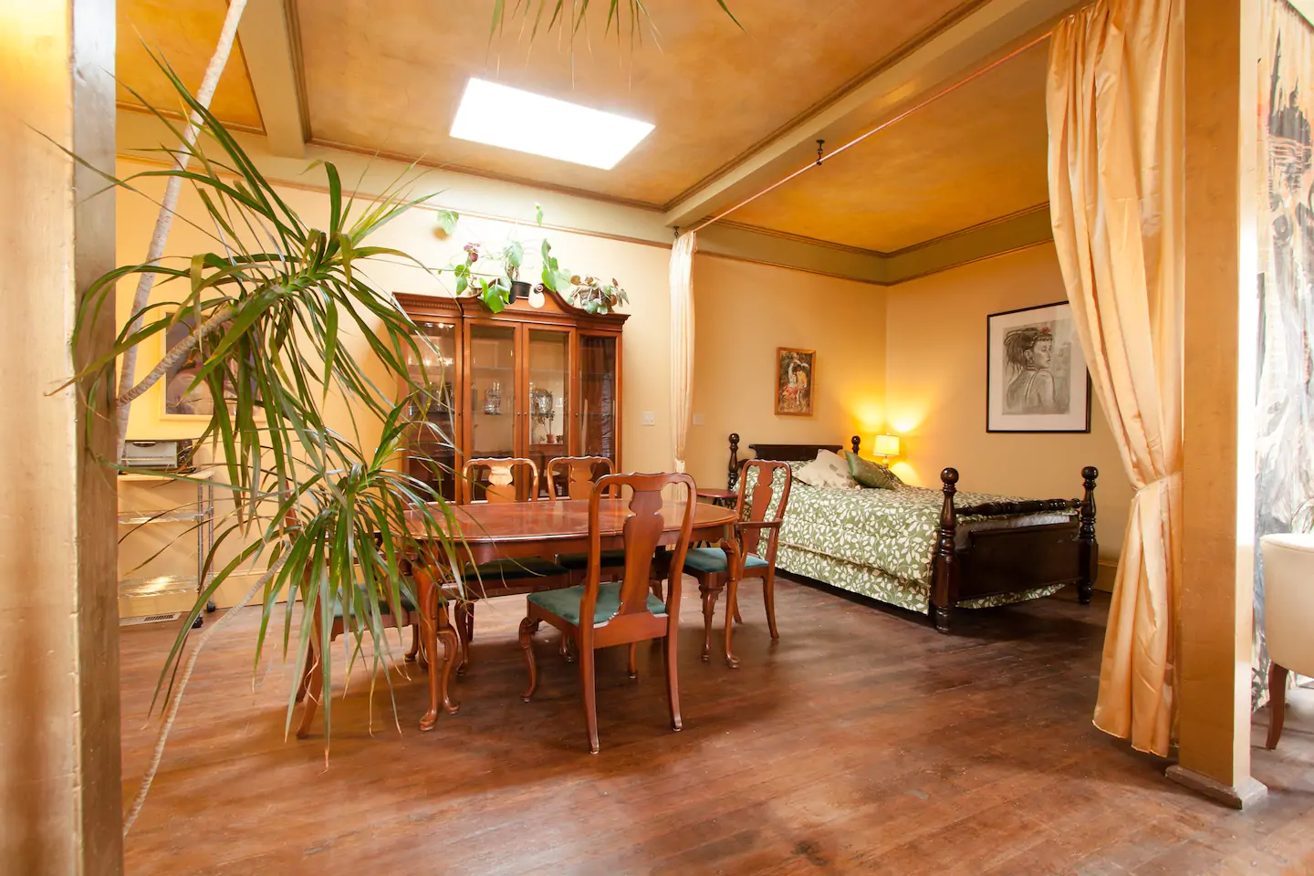 Warm wooden dining room table with chairs all around it. In the far back corner is a bed with green bedding.