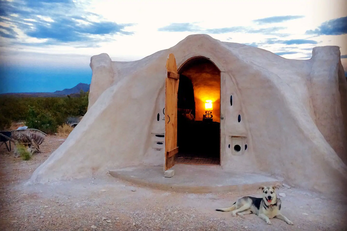 A khaki-colored dome building has a door open, leading to a room. A dog sits outside the home.