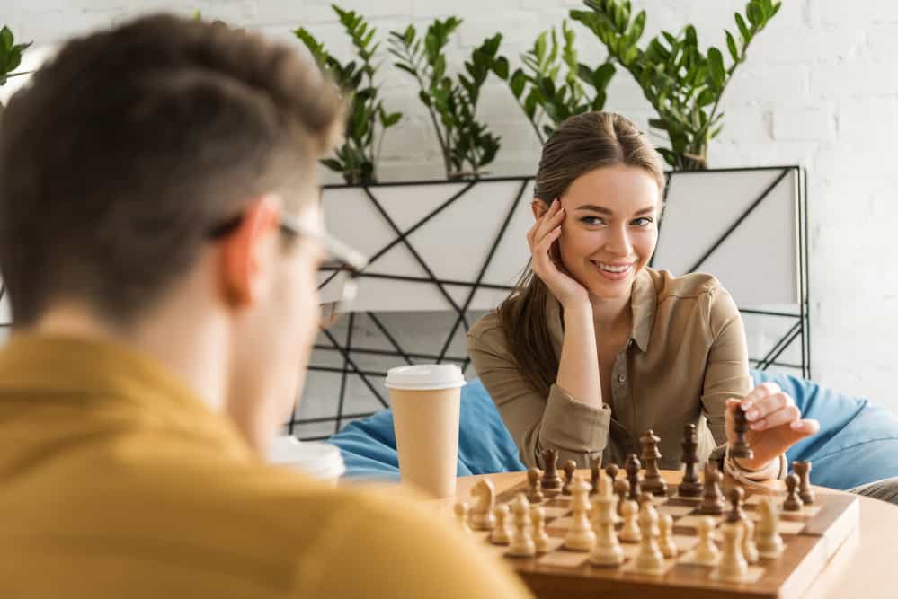 A girl smiles at a man across the table. A coffee cup and a chess board sits on the table between them.