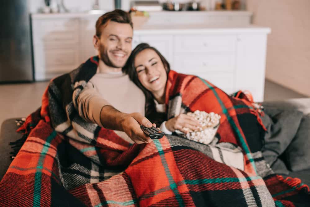 A man and woman snuggle in a blanket with popcorn.