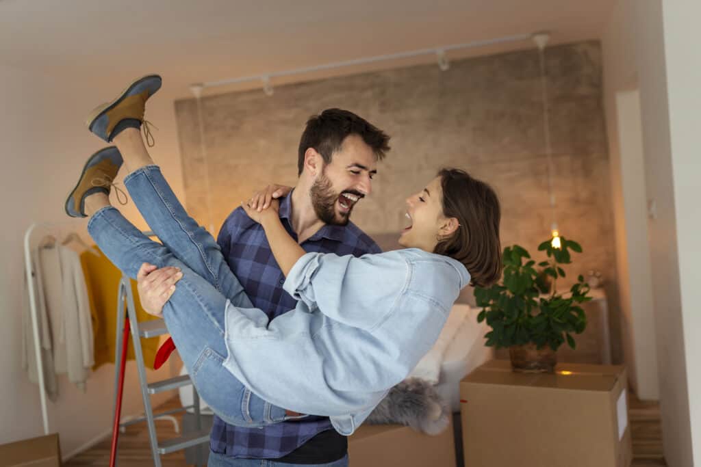 Beautiful newly married young couple moving in together, having fun while unpacking cardboard boxes with their belongings, husband carrying wife in his arms